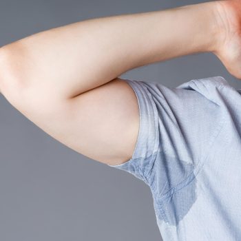Shirt displays sweat stain under the arm while the person's arm is raised, set against a plain gray background.