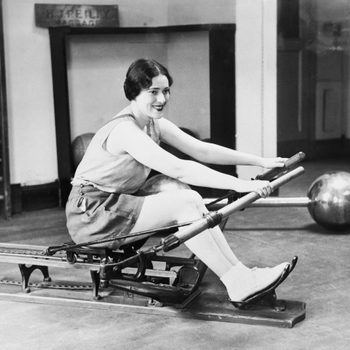 A woman exercises on a vintage rowing machine indoors, surrounded by gym equipment including a large exercise ball.