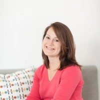 A person smiles, seated on a sofa, against a white wall with a patterned cushion nearby.