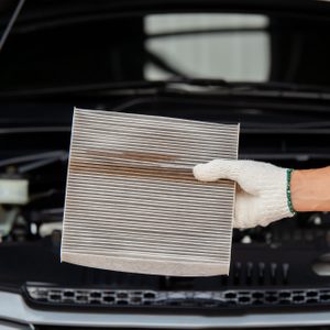 A hand in a glove holds a used car air filter, displayed in front of an open car hood.