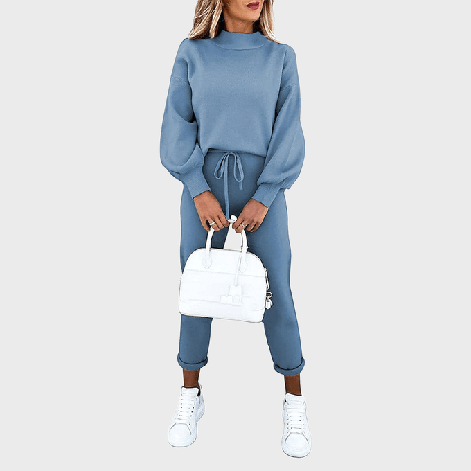 Person stands holding a white handbag, wearing a blue sweater and pants, paired with white sneakers, against a plain background.