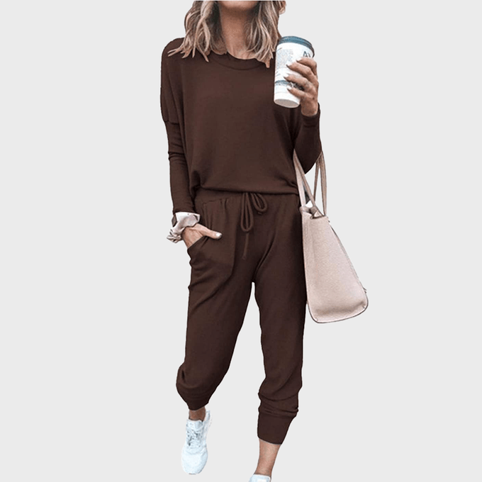 A person wearing a brown tracksuit holds a coffee cup and beige bag, standing against a plain background.