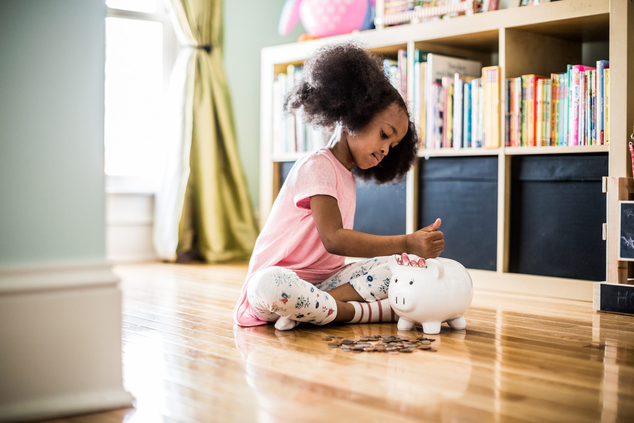 A child places coins into a piggy bank on a hardwood floor, surrounded by a bookshelf and soft natural light.