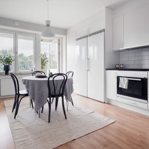 Table covered with gray cloth sits on rug; chairs surround it in a modern kitchen with white cabinets and large windows.