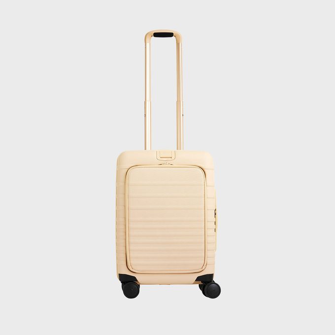 A beige suitcase with an extended handle stands upright on four black wheels against a plain gray background.