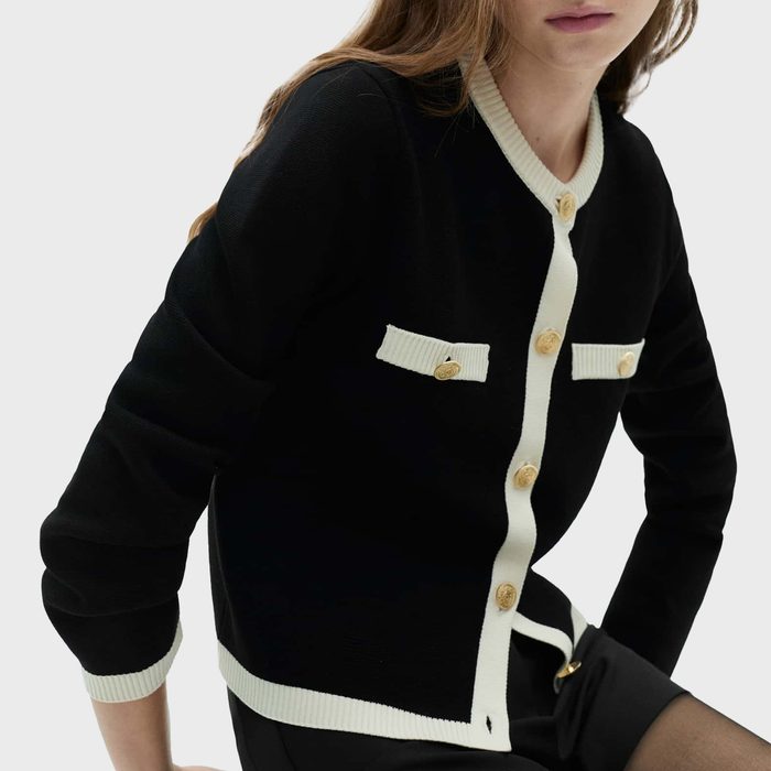 A woman with long, wavy hair wears a black cardigan with a round neckline, white trim, long sleeves, and gold buttons, paired with a black skirt